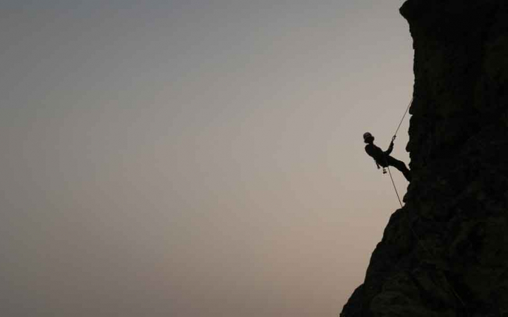 the silhouette of a rock climber resting midway up a rock wall